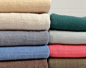 May include: A stack of folded fabric swatches in various colors including beige, cream, gray, blue, brown, dark green, light green, and coral. Each piece of fabric features a visible, loose-weave, textured surface with a consistent grid-like pattern.
