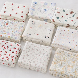 May include: A collection of nine folded, white muslin cloths with various patterns. Designs include strawberries, cherries, floral arrangements, polka dots, and cartoon animals. The cloths are stacked neatly, showcasing the variety of patterns.