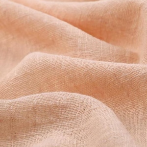 May include: A soft, light peach-colored fabric with a visible woven texture. The material is draped in gentle folds, creating a series of soft shadows and highlights across the surface that emphasize the loose, breathable weave of the textile.