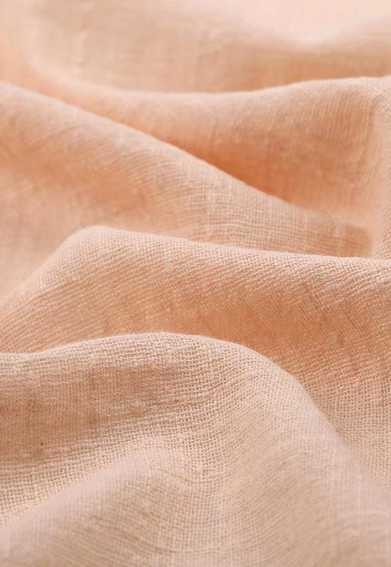 May include: A soft, light peach-colored fabric with a visible woven texture. The material is draped in gentle folds, creating a series of soft shadows and highlights across the surface that emphasize the loose, breathable weave of the textile.