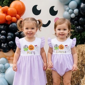 May include: Two children wearing matching white tops with Halloween graphics and light purple gingham bottoms. One child wears a dress, the other a romper. Both tops feature the names Mary and Enna in glittery text, set against a balloon arch.
