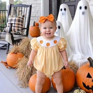 May include: A light yellow baby romper with short puff sleeves and a white smocked collar featuring embroidered orange pumpkins. The outfit is paired with a matching orange hair bow and brown leather shoes, creating a festive seasonal look.