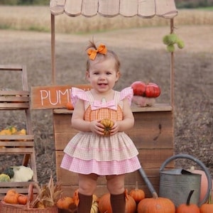 Puede incluir: Un niño pequeño viste un vestido sin mangas con un corpiño naranja con estampado de calabazas y una falda de capas en rosa y beige con cuadros vichy. El conjunto incluye un lazo naranja a juego para el pelo y calcetines marrones altos.