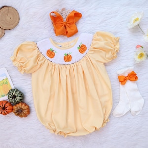 May include: A pale yellow bubble romper featuring a white smocked collar with embroidered orange pumpkins, short puff sleeves, a matching orange hair bow, and white knee-high socks with orange bows, presented as a complete seasonal outfit set.