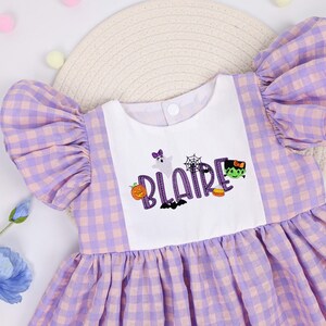 May include: A dress featuring a white bodice with the name BLAIRE embroidered in purple letters, surrounded by Halloween icons including a ghost, spiderweb, pumpkin, bat, candy corn, and Frankenstein's monster, with purple gingham sleeves and skirt.