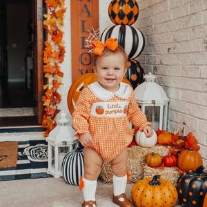 May include: An orange and white gingham long-sleeve romper featuring a white collar with embroidered text and a central smocked panel with the text little pumpkin. The outfit includes white knee-high socks with orange bows and brown leather shoes.