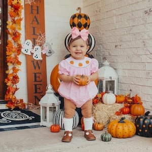 May include: A light pink bubble romper featuring a smocked neckline with embroidered orange pumpkins, paired with white knee-high socks and brown leather shoes. A large pink hair bow is worn on the head, and a small orange pumpkin is held in the hands.