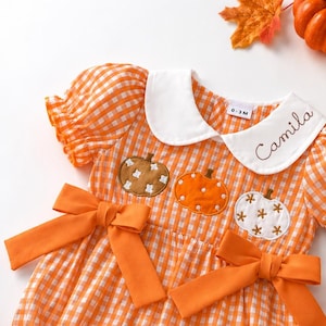 May include: An orange and white gingham dress with short puff sleeves, a white Peter Pan collar embroidered with the name Camila, and two orange waist bows. The bodice features three embroidered pumpkin appliques in brown, orange, and white.