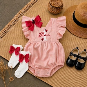 May include: A light pink romper with ruffled sleeves, featuring a red bow at the neckline and the embroidered name Blair with a number one and ladybug graphic. Includes white ribbed socks with red bows and black patent leather mary jane shoes.