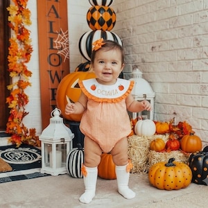 May include: A baby wearing an orange and white gingham romper with a ruffled collar featuring embroidered pumpkins and the name Octavia, paired with white knee-high socks with orange bows and a matching orange hair bow, standing in a festive setting.