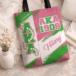 Puede incluir: Bolso tote rosa y verde con asas negras. La bolsa presenta el texto "AKA 1908" en verde y rosa, junto con el nombre "Hilary" en cursiva. El diseño incluye rayas de pinceladas verdes y rosas y las letras "AKA" en verde.
