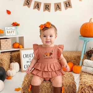 May include: A rust-coloured ruffled romper featuring an embroidered pumpkin design and the name Sarah on the chest. The outfit includes a matching orange hair bow and brown ribbed knee-high socks with small orange bows attached to the sides.