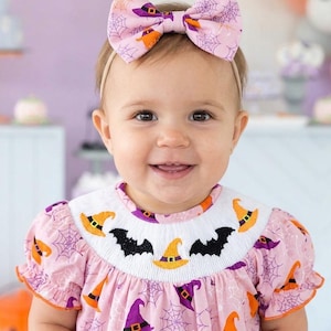 May include: A light pink dress featuring a smocked bodice with embroidered black bats and orange witch hats, paired with a matching hair bow. The fabric has a repeating pattern of purple spiderwebs, orange witch hats, and purple witch hats.