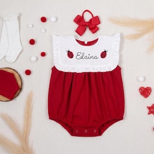 Può includere: Una tutina rossa e bianca con colletto a balze, due illustrazioni di coccinelle e il nome Elaina ricamato in corsivo nero. Il set include un paio di calzini bianchi, un fiocco rosso per capelli e un quadrato di tessuto rosso su base in legno.