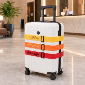 May include: A white rolling suitcase with a black handle and wheels. The suitcase has three colorful straps with the names "Riley", "Evelyn", and "Brooklyn" printed on them. The straps are yellow, orange, and red.