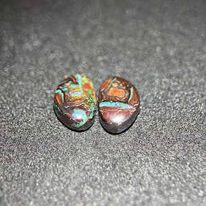 May include: Two oval-shaped gemstones with a dark brown base color. The stones have intricate patterns of orange, teal, and green, with hints of blue and purple. The gemstones are set against a dark, textured background.