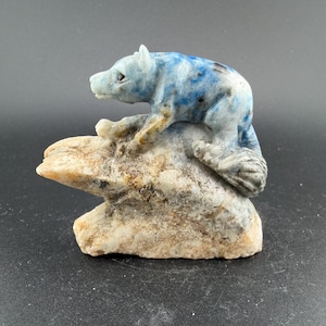 May include: A carved stone sculpture of a bear in shades of blue and white, with dark blue speckles, perched atop a light brown rock formation. The bear is detailed with visible paws and a defined snout.