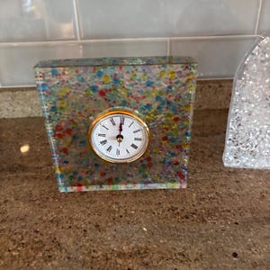 May include: A square decorative clock with a gold-tone bezel and a white clock face. The clock is encased in a clear resin with colorful specks. The clock is on a brown surface.