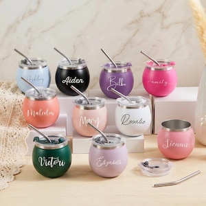 Puede incluir: Una colección de vasos de acero inoxidable personalizados y coloridos con tapas y pajitas. Cada vaso es de un color diferente, con nombres como "Ashley" y "Aiden" en escritura cursiva. Un jarrón con hierba de la pampa y pequeñas flores blancas en el fondo.