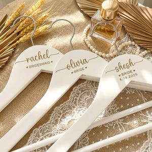Puede incluir: Tres perchas de madera blanca con ganchos plateados, personalizadas con nombres y títulos en escritura dorada. Una dice "Rachel - Bridesmaid", otra "Olivia - Bride" y la tercera "Shesh - Bride 4.19.2025". Las perchas están dispuestas sobre una superficie texturizada.