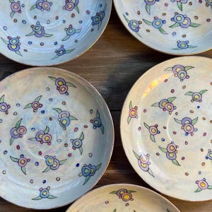 Peut inclure: Ensemble de six assiettes décoratives en céramique. Chaque assiette présente une base crème avec un éclat nacré et est ornée d'un motif floral stylisé répété dans des couleurs pastel, notamment le bleu, le violet et le rose. Les assiettes ont un bord doré.