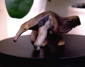 May include: A small, detailed anteater figurine featuring a long, slender snout, a black stripe along its side, and textured brown fur. The animal is depicted in a walking pose with its head lowered, showcasing realistic anatomical features.