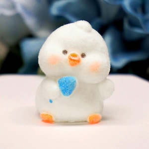 May include: A fluffy white chick figurine with a blue heart-shaped object. The chick has orange feet and beak, rosy cheeks, and small black eyes. The figurine is a cute, whimsical design.