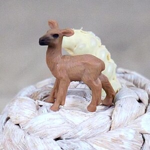 May include: A small, brown deer figurine with a dark muzzle and ears stands on a white, woven surface. The deer is positioned in front of a light yellow, textured object. The figurine is a miniature representation of a deer.
