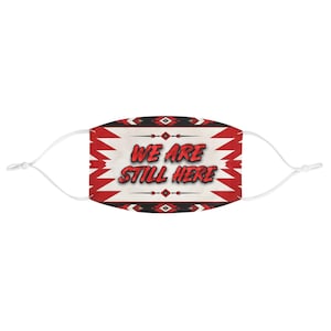 Op de afbeelding: Een gezichtsmasker met de tekst WE ARE STILL HERE in dikke rode letters. Het ontwerp heeft een witte achtergrond met rode en zwarte geometrische patronen langs de boven- en onderrand, en witte elastische oorlussen met verstelbare knopjes.