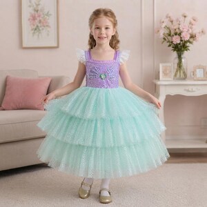 May include: A floor-length dress featuring a sequined purple bodice with a shell-shaped accent at the center and a multi-layered, light green tulle skirt with a shimmering, grid-like pattern. The dress includes short, ruffled white tulle sleeves.