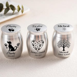 May include: Three silver-toned metal urns with cylindrical bodies and rounded lids. Each urn features black laser-engraved designs: the first shows a dog silhouette and the name Harley, the second a heart of paw prints and James Smith, and the third a tree and Micheal.