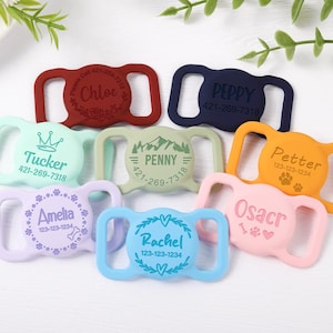 May include: A collection of eight silicone pet collar tags in various colors including red, navy, mint, sage, orange, lavender, light blue, and pink. Each rectangular tag features a central circular area with engraved names and phone numbers.