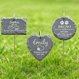May include: Three slate memorial garden stakes in grass. Each stake has a different shape and features engraved names, dates, and paw prints. One is rectangular, one is heart-shaped, and one is circular.