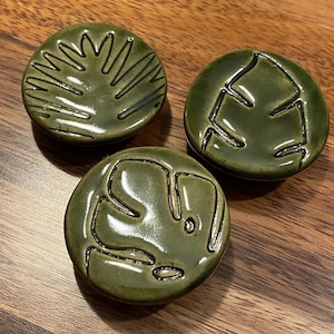 May include: Three circular ceramic buttons in a glossy olive green glaze. Each button features a distinct incised botanical pattern, including a palm-like leaf design and two variations of monstera leaf motifs with dark, recessed lines.
