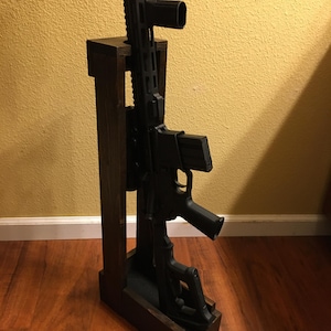 Può includere: Un fucile nero è esposto in un supporto per fucile in legno marrone scuro. Il supporto è rettangolare con un supporto verticale e una base. Il fucile è posizionato verticalmente, mostrando il suo design e le sue caratteristiche.