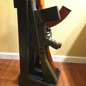 May include: A black wooden gun rack holding two firearms. One gun is olive green and black, the other is brown. The rack is rectangular and sits on a wooden floor.