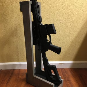 May include: A black rifle is displayed in a gray wooden gun rack. The rack has a rectangular base and two vertical supports. The rifle is positioned upright, showcasing its details against a yellow wall and a wooden floor.