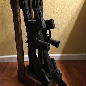 Pode incluir: Um suporte de madeira castanho escuro com várias armas de fogo pretas. O suporte tem uma base retangular e é tingido de castanho escuro. As armas de fogo são presas com correias pretas. O fundo é uma parede amarelo claro e um chão de madeira.