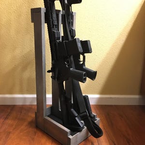 May include: A gray wooden gun rack holding multiple black rifles. The rack has a rectangular base and a vertical support structure. The rifles are positioned upright, with their barrels facing upwards. The rack is on a wooden floor.
