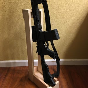 May include: A black rifle with a black strap is displayed on a light-coloured wooden stand. The stand is rectangular with a base and a vertical support. The background is a light yellow wall and a wooden floor.