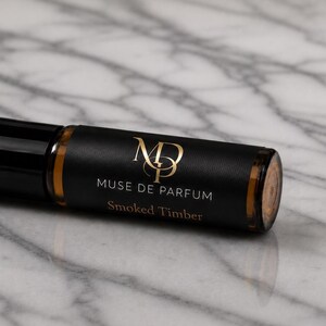 May include: A roll-on perfume bottle with a black cap and a black label with gold lettering. The label reads "Muse de Parfum" and "Smoked Timber". The bottle is on a marble surface.
