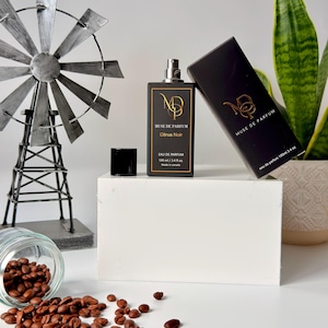 K&ouml;nnte beinhalten: Eine schwarze Parf&uuml;mflasche und -schachtel mit goldfarbenen Buchstaben, die "MUSE DE PARFUM Citrus Noir" lauten. Die Flasche befindet sich neben einer schwarzen Kappe und einer dekorativen Windm&uuml;hle. Kaffeebohnen sind auf der wei&szlig;en Oberfl&auml;che verstreut.