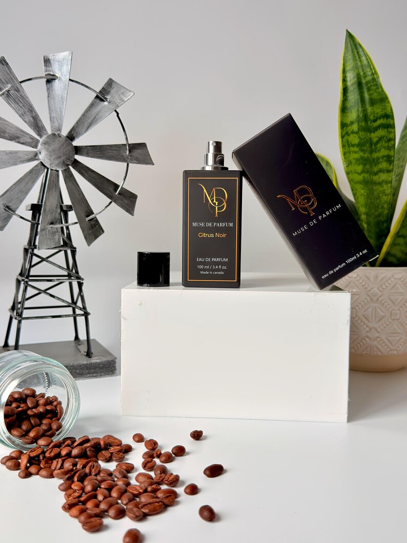 K&ouml;nnte beinhalten: Eine schwarze Parf&uuml;mflasche und -schachtel mit goldfarbenen Buchstaben, die "MUSE DE PARFUM Citrus Noir" lauten. Die Flasche befindet sich neben einer schwarzen Kappe und einer dekorativen Windm&uuml;hle. Kaffeebohnen sind auf der wei&szlig;en Oberfl&auml;che verstreut.