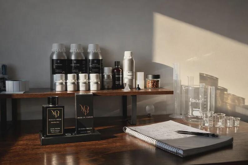 May include: A perfume-making workspace with various bottles, jars, and beakers. Several silver bottles and small glass jars sit on a wooden shelf. Two perfume bottles and boxes are on a black stand. A notebook and pen are on the table.