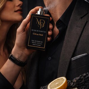 K&ouml;nnte beinhalten: Schwarze rechteckige Parf&uuml;mflasche mit goldfarbenem Schriftzug und dem Text "MUSE DE PARFUM Citrus Noir". Die Flasche wird auf den Hals einer Person gespr&uuml;ht. Eine Zitronenscheibe befindet sich im Vordergrund. Die Flasche enth&auml;lt 100 ml.