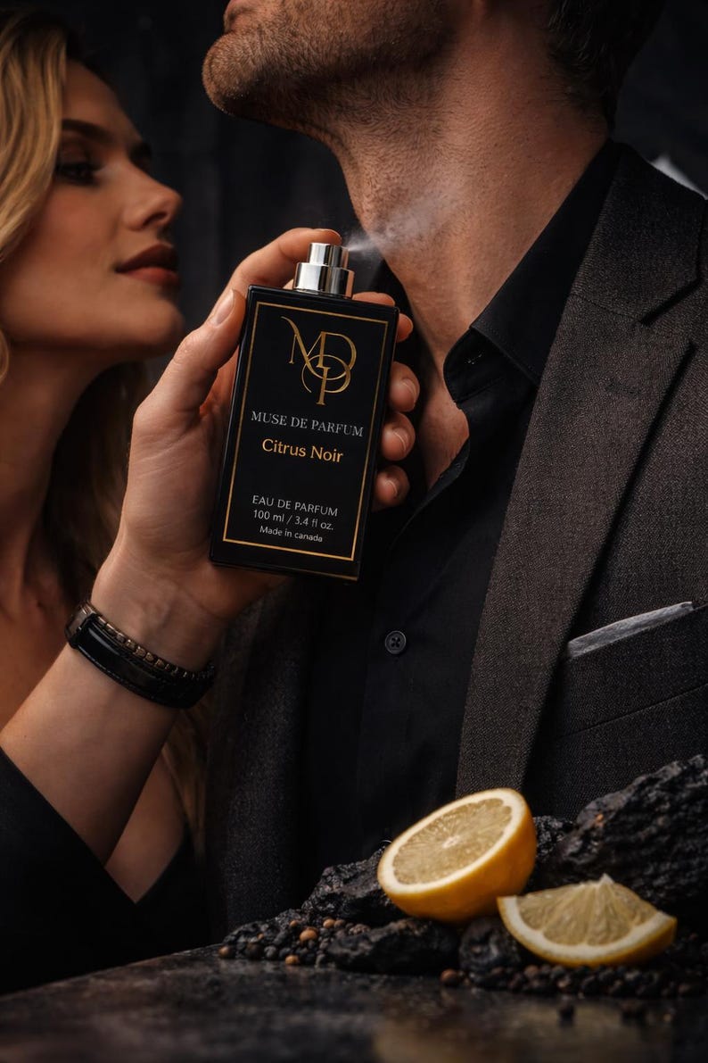 K&ouml;nnte beinhalten: Schwarze rechteckige Parf&uuml;mflasche mit goldfarbenem Schriftzug und dem Text "MUSE DE PARFUM Citrus Noir". Die Flasche wird auf den Hals einer Person gespr&uuml;ht. Eine Zitronenscheibe befindet sich im Vordergrund. Die Flasche enth&auml;lt 100 ml.
