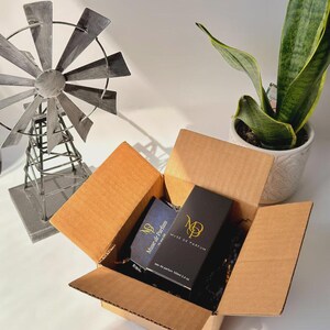 May include: An open cardboard box containing two perfume boxes. One box is black with gold lettering and the other is blue with gold lettering. The boxes are labeled "Muse de Parfum". A decorative windmill and a potted plant are in the background.