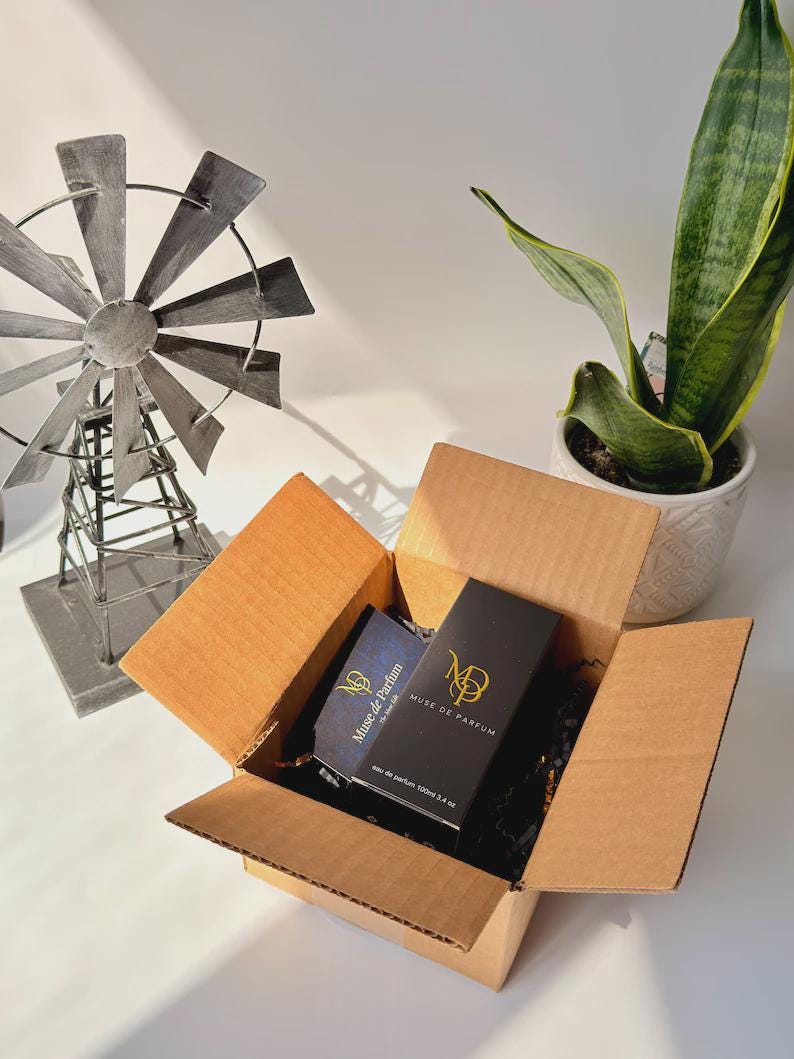 May include: An open cardboard box containing two perfume boxes. One box is black with gold lettering and the other is blue with gold lettering. The boxes are labeled "Muse de Parfum". A decorative windmill and a potted plant are in the background.