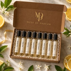 May include: A brown cardboard gift box containing eight clear glass perfume roller bottles with black caps. Each bottle features a white label with the text Midnight Oud, Amber Drift, Cedar Noir, Ocean Breeze, Spiced Tobacco, Vetiver Smoke, Leather Essence, and Black Amber.