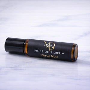 K&ouml;nnte beinhalten: Eine bernsteinfarbene Roll-on-Parf&uuml;mflasche mit schwarzem Verschluss und dunklem Etikett. Das Etikett tr&auml;gt den goldenen Text "MUSE DE PARFUM Citrus Noir". Die Flasche liegt auf einer wei&szlig;en Marmoroberfl&auml;che.
